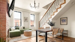 A modern living room with a green sofa, a dining table with chairs, a staircase, and large windows.