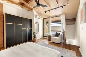 Une chambre à coucher comprenant un lit, un fauteuil blanc, une grande armoire, un ventilateur de plafond et une vue sur un balcon.