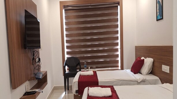 A hotel room with two beds, a TV mounted on the wall, a chair, and a window with blinds. - Hotel Faaz International (Kannur)