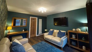 A living room with a beige sofa, a white sofa, a blue rug, a wooden side table, a TV mounted on the wall, and a door with a window. - Cochrane House (Stirling)