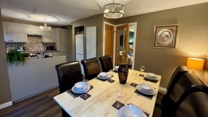 A dining area with a table set for four, surrounded by chairs, in a modern kitchen with white cabinets and a tiled backsplash. - Cochrane House (Stirling)