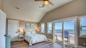 Une chambre avec un lit, une table de chevet, un ventilateur de plafond et une vue sur la plage grâce à de grandes portes-fenêtres coulissantes.