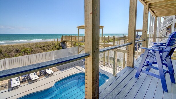 Une terrasse en bois avec une piscine, des chaises longues et une vue sur la plage et l’océan.