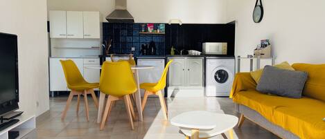 A modern living room with a sofa, a dining table with chairs, a TV, and a kitchen area with a washing machine.
