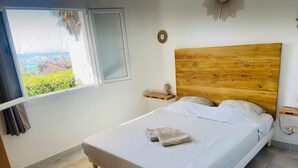 A bedroom with a wooden headboard, a bed with patterned bedding, a nightstand with a lamp, and a view of greenery through the window. - Apartment T2 furnished tourism 2 * air-conditioned near the sea (Olmeto)
