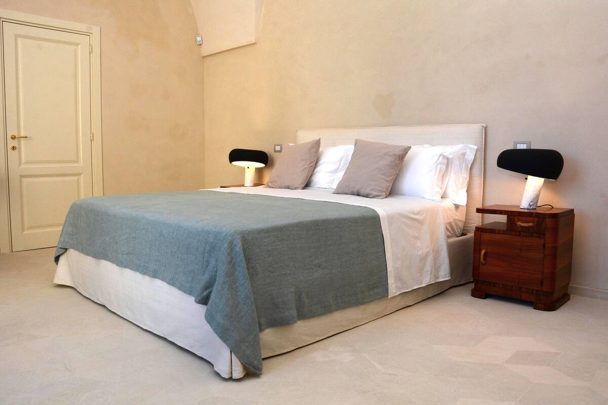Una cama bien hecha con una colcha gris y blanca, dos lámparas de noche y una mesita de noche de madera con un cajón.