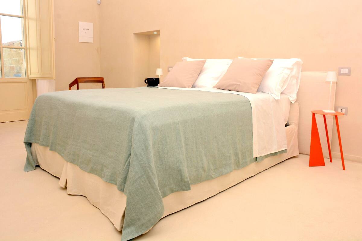 Una cama bien hecha con una colcha azul claro y almohadas blancas, una pequeña silla de madera y un taburete rojo al lado de la cama.