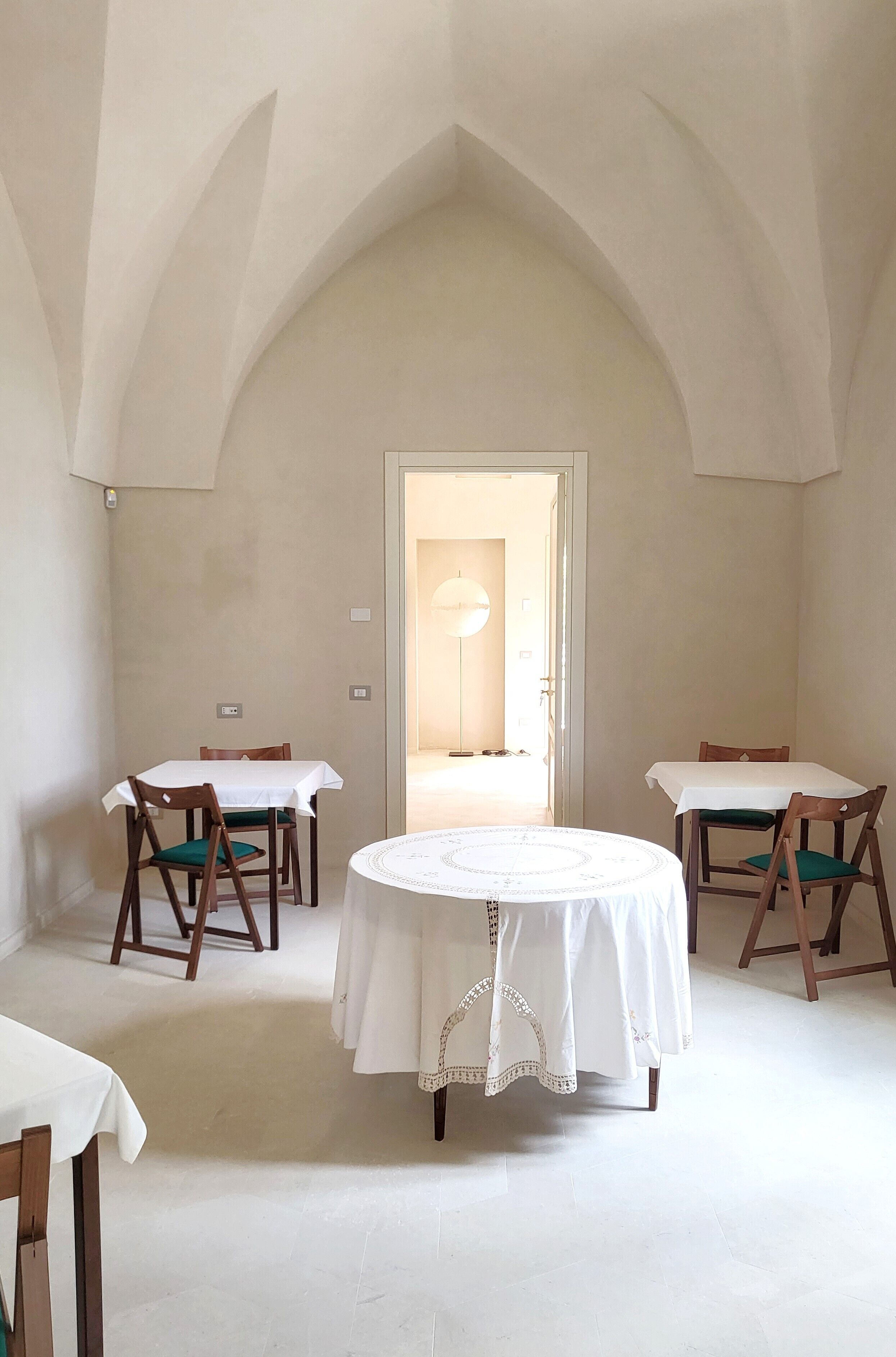 Un comedor con dos mesas, cada una con mantel blanco y sillas de madera. La habitación tiene un techo alto en arco y una puerta que da a otra estancia.