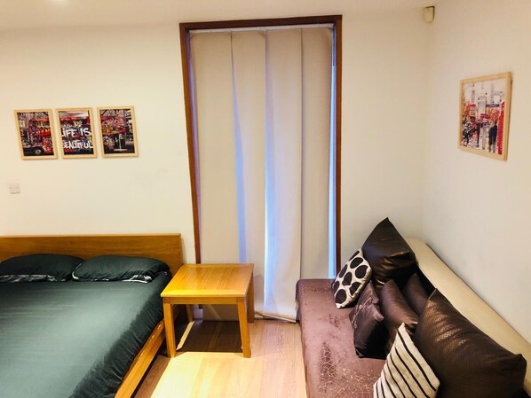 A small, furnished room with a bed, a sofa, a small table, and a window with curtains. There are three framed pictures on the wall.