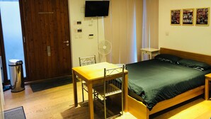 A bedroom with a bed, a desk, a chair, a TV, and a fan.