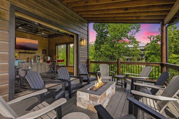 Eine Terrasse mit Adirondack-Stühlen, einer Feuerstelle und Blick auf Bäume und einen Sonnenuntergang.