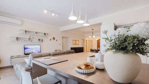 A modern kitchen with a central island, bar stools, a dining area, and a living room with a sofa and TV.