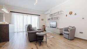 A modern living room with a sofa, armchair, and a small round table. There is a TV on a white cabinet, and a large window with sheer curtains.