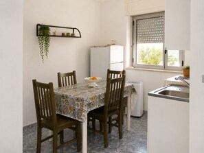 Een compacte keuken met een eettafel, stoelen, een koelkast, een spoelbak en een raam met zonwering.