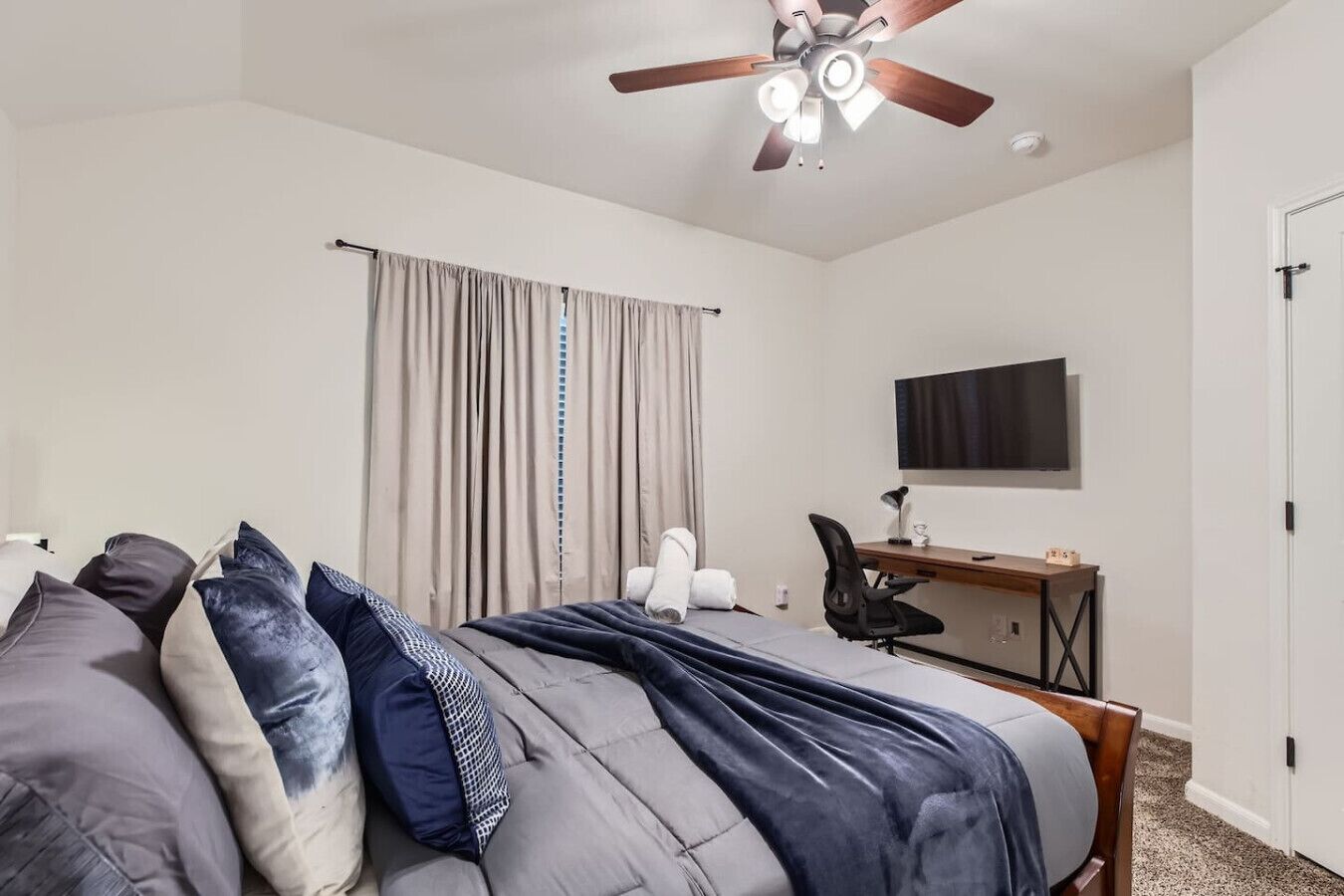 Quarto com cama, escrivaninha, cadeira, ventilador de teto e televisão na parede.