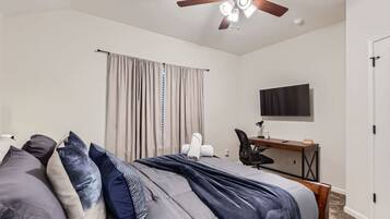 Quarto com cama, escrivaninha, cadeira, ventilador de teto e televisão na parede.