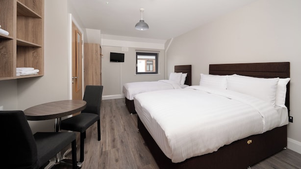 Habitación de hotel con dos camas, un escritorio y una silla. Se observa un armario con estantes y un televisor fijado a la pared.