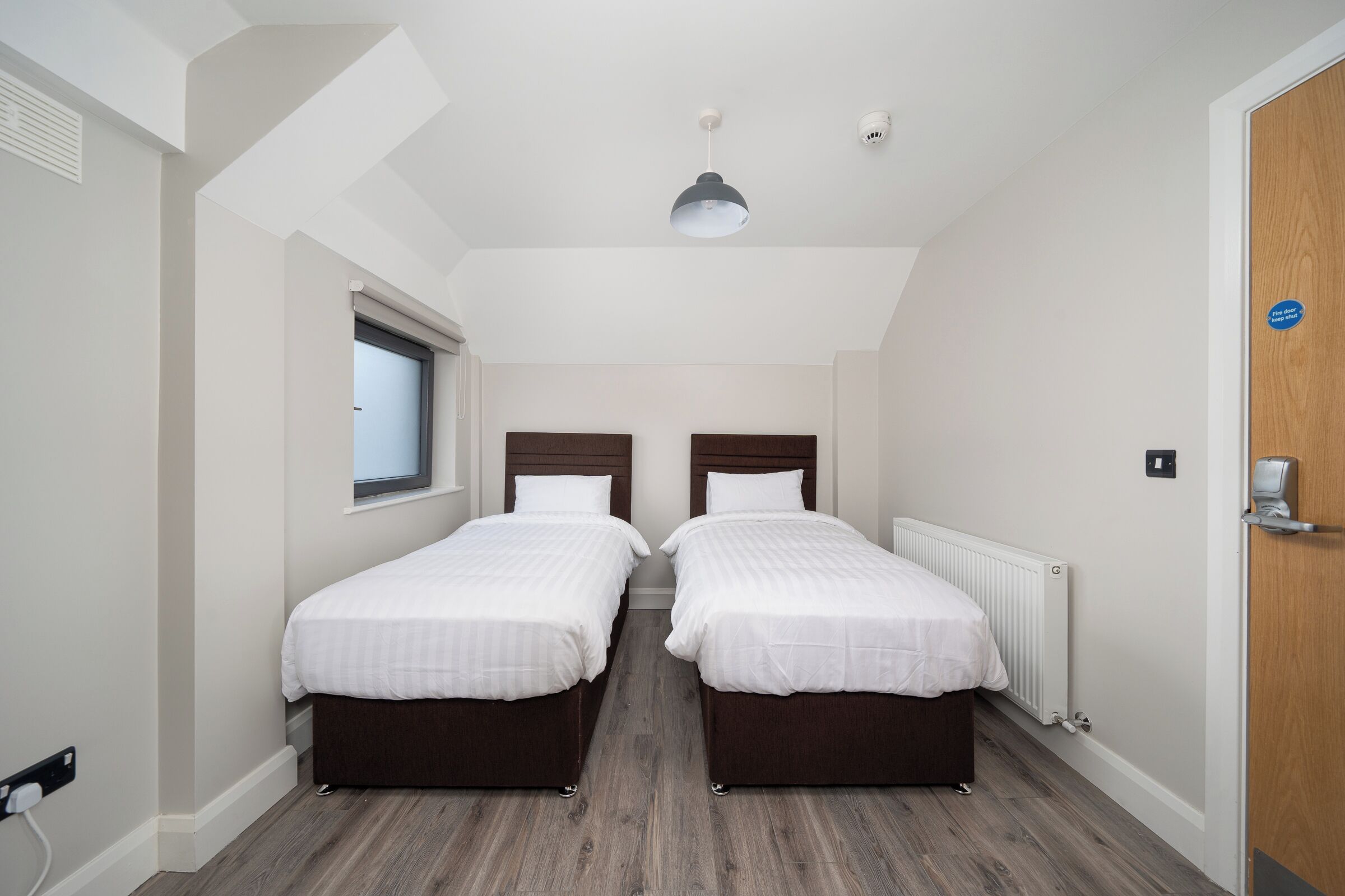 A room with two single beds, a window, a radiator, and a door with a sign.