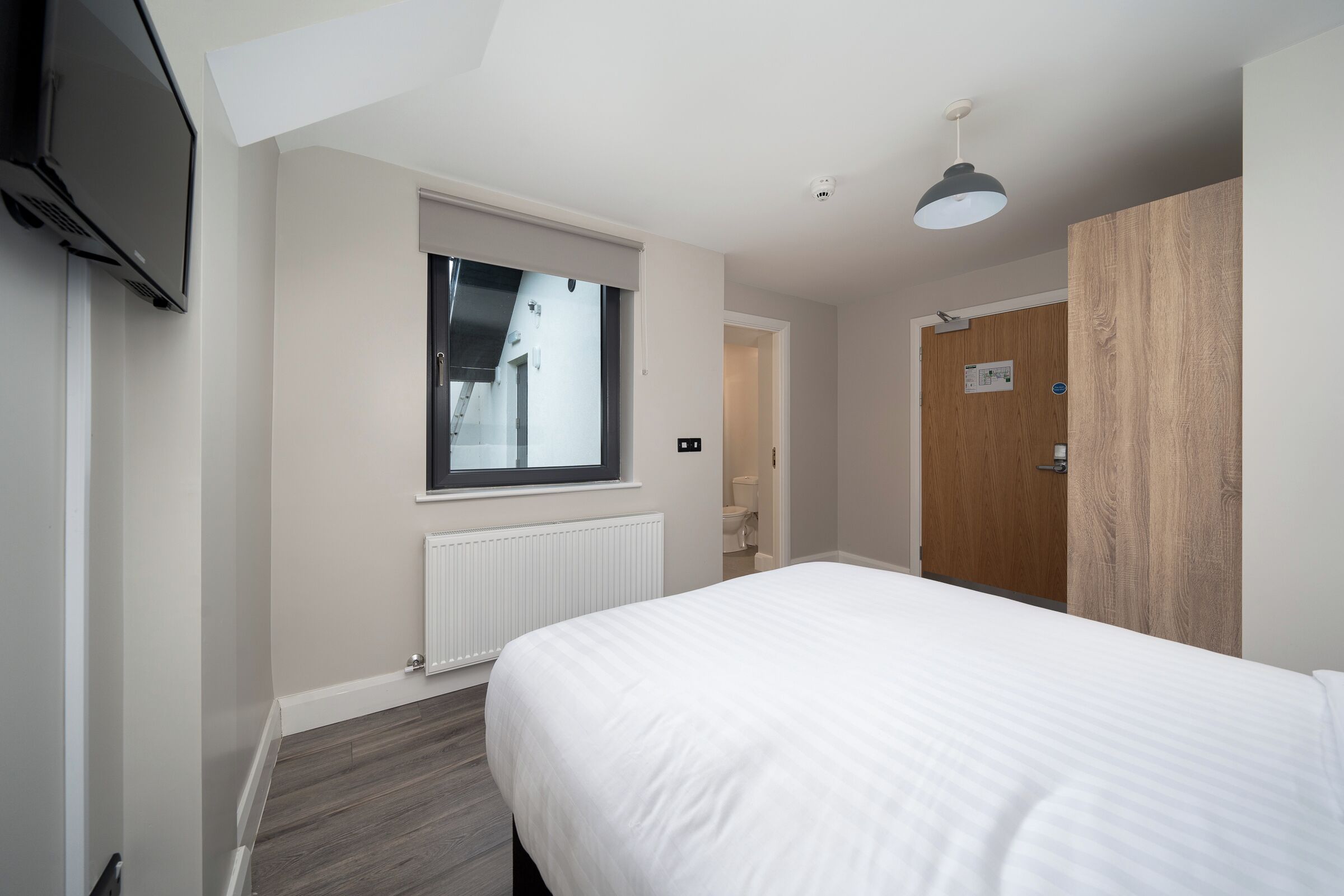 A modern hotel room with a large bed, a window, a radiator, a wooden wardrobe, and a television mounted on the wall.