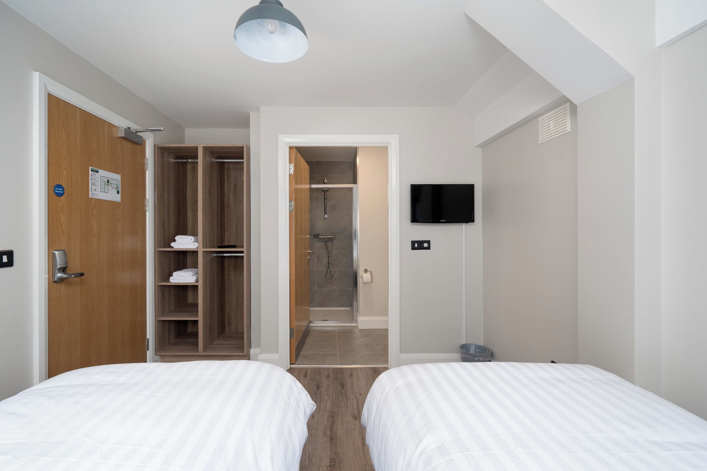 Habitación de hotel con dos camas, un armario de madera, zona de ducha y un televisor en la pared.
