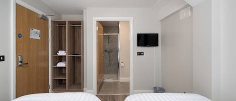 A hotel room with two beds, a wooden wardrobe, a shower area, and a television on the wall.
