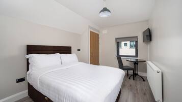 A neatly made bed with white linens, a small round table with a chair, a television mounted on the wall, and a window with a view of buildings.