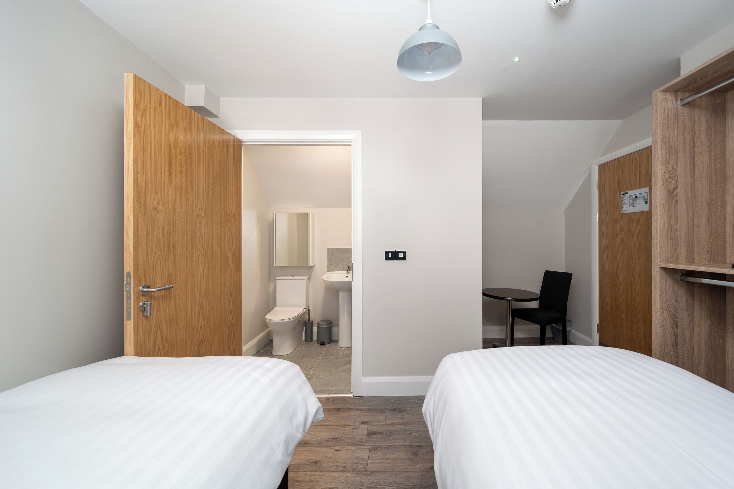 A hotel room with two beds, a wooden wardrobe, a desk, and a bathroom visible through an open door.