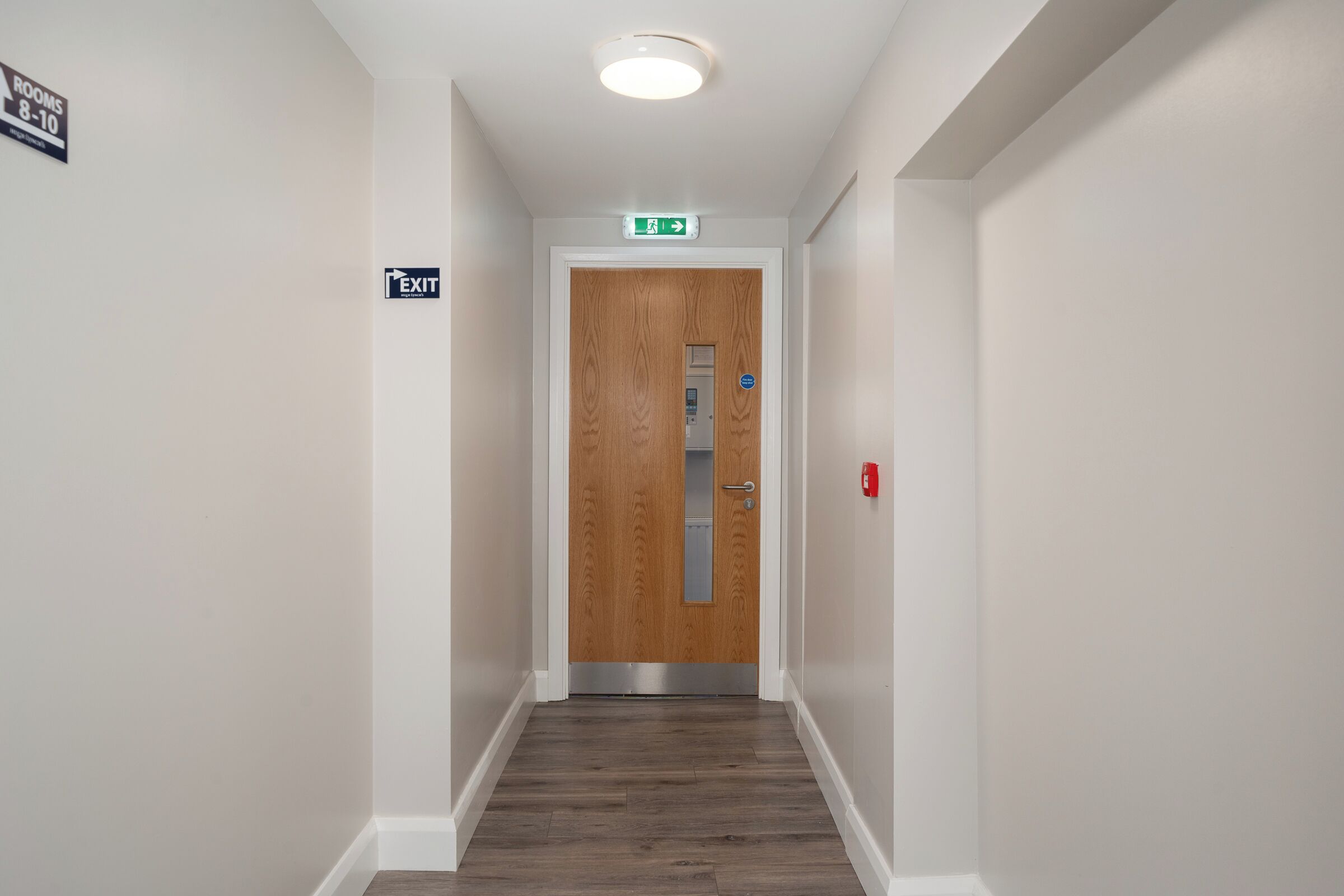 A hallway with a wooden door, an exit sign, and a fire alarm.