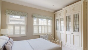 A bedroom with a bed, two windows with shutters, a radiator, and a wardrobe with glass doors. - Kings Road 3 bed House Chelsea (London)