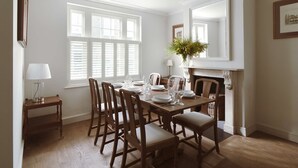 A dining room with a wooden table set for six, flanked by white walls and large windows with shutters. - Kings Road 3 bed House Chelsea (London)