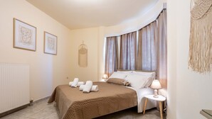 A neatly arranged bedroom with a bed, two bedside lamps, a wall-mounted macrame piece, and two framed artworks. - Villa Amare - Heraklion Private Garden Retreat (Hersonissos)