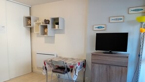 Ein kleiner, ordentlicher Raum mit einem Tisch, einem Stuhl, einem Fernseher auf einer Holzkommode und einigen Regalen an der Wand mit Dekoration.