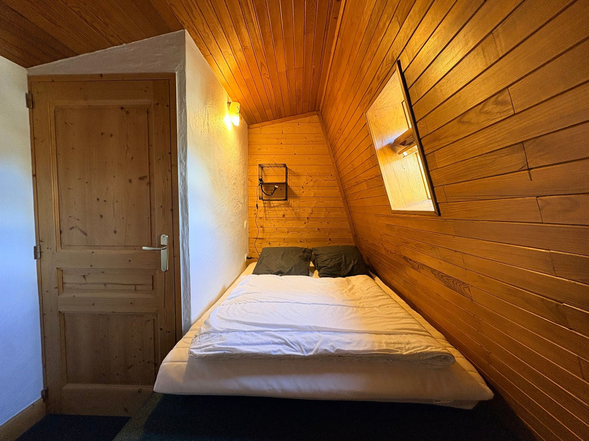 A bedroom with a wooden sloped ceiling, a single bed, a door, and a small window.