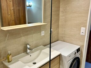 A bathroom with a white sink, a mirror, a glass partition, a bottle of liquid soap, and a washing machine.