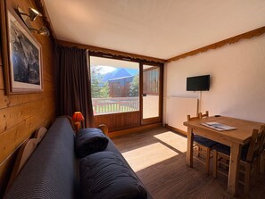 A room with a wooden bench, a table with chairs, a television, and a window with a view of buildings and trees. - Bright studio, ski-in ski-out, pets allowed (Les Deux Alpes)