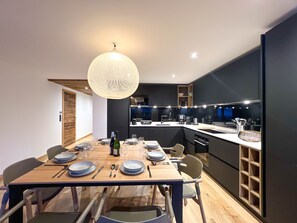 A modern kitchen with a dining area, featuring a wooden table set for four, a hanging light fixture, and built-in appliances.