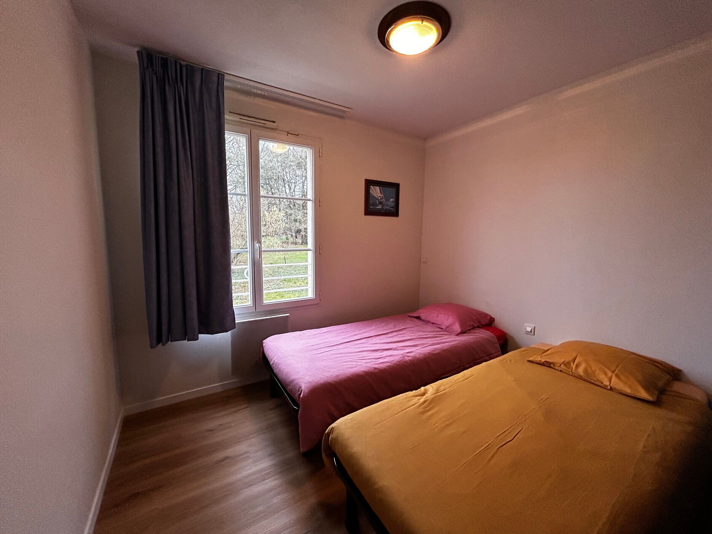 A small, single-bed room with a window, a picture on the wall, and a ceiling light.