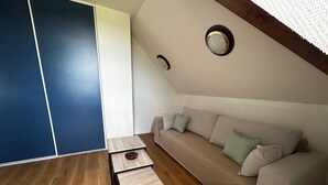 A room with a wooden floor, a beige sofa with cushions, a small wooden table, and a ceiling light fixture. - Renovated T2 with heated pool near Connétable Spa, parking and WIFI (La Roche-Posay)