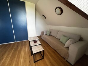 A room with a wooden floor, a beige sofa with cushions, a small wooden table, and a ceiling light fixture. - Renovated T2 with heated pool near Connétable Spa, parking and WIFI (La Roche-Posay)