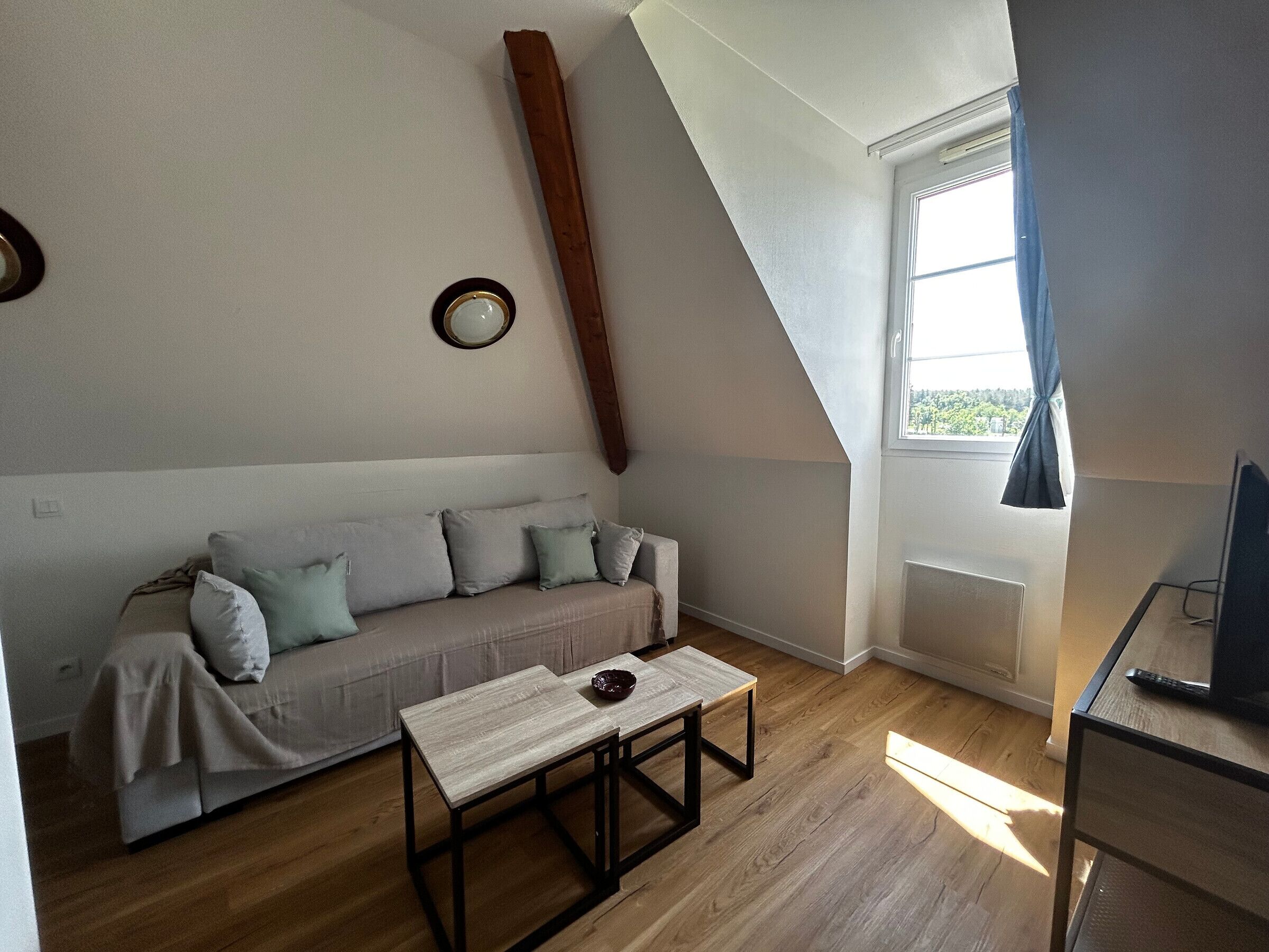 Une pièce avec un plafond en pente, un canapé avec des coussins, une petite table et une fenêtre avec des rideaux.
