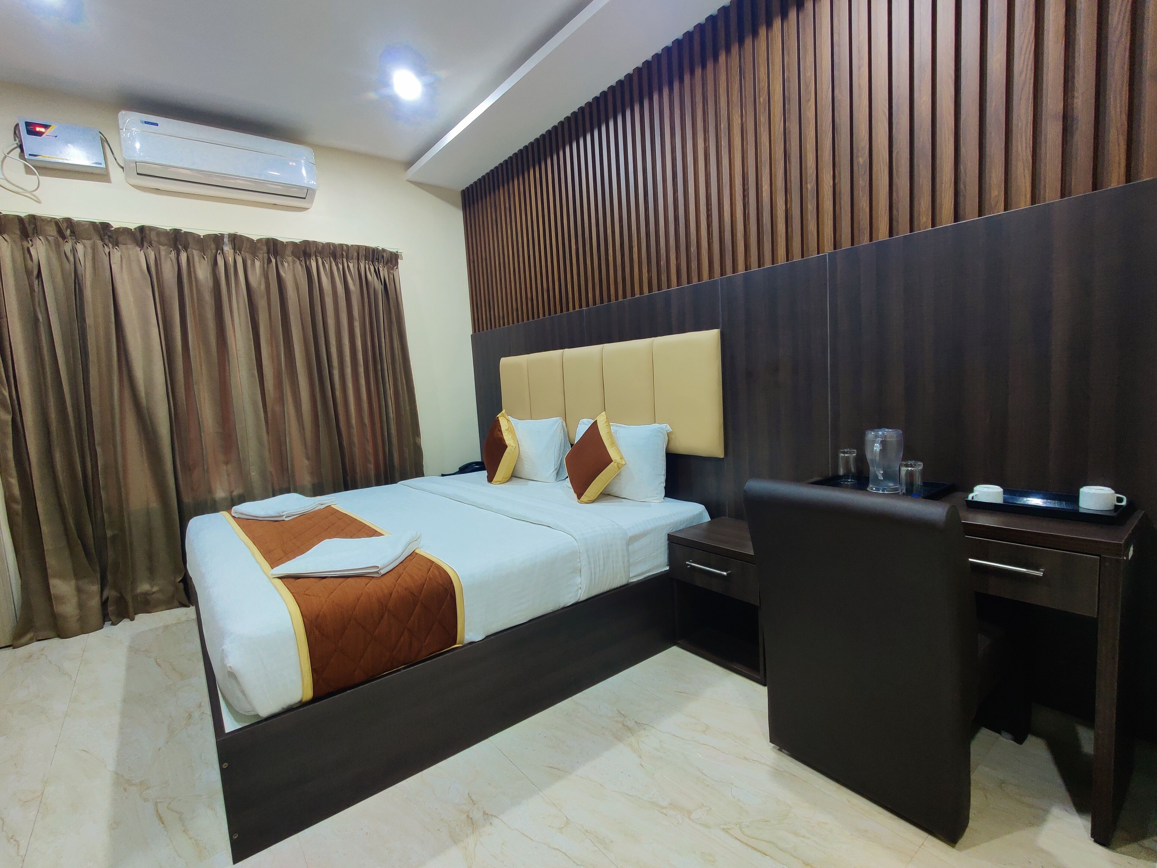 A hotel room with a bed, bedside table, desk, and chair. There are air conditioning units on the wall, a curtain, and a wooden panel wall.