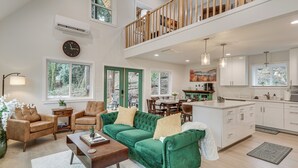 A spacious living area with a green sofa, wooden chairs, and a coffee table. A kitchen with white cabinets and a dining area with a table and chairs are visible. - Near Rivers & Boating: Home w/ Hot Tub in Longview (Longview)
