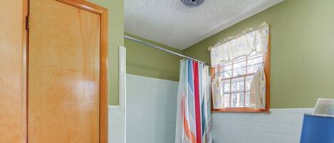 A bathroom with a wooden door, a bathtub, a shower curtain, a window with a curtain, a sink, and a trash can.