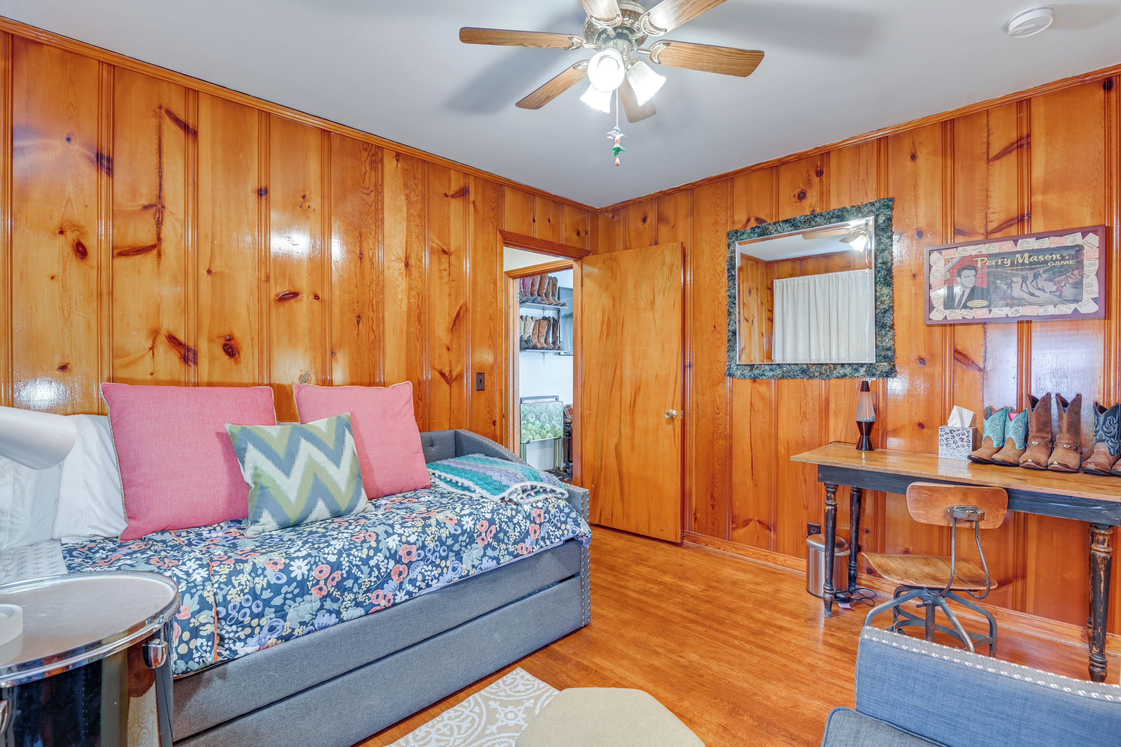 A cozy room with wooden walls, a bed with colorful bedding, a desk with a mirror, and a ceiling fan.