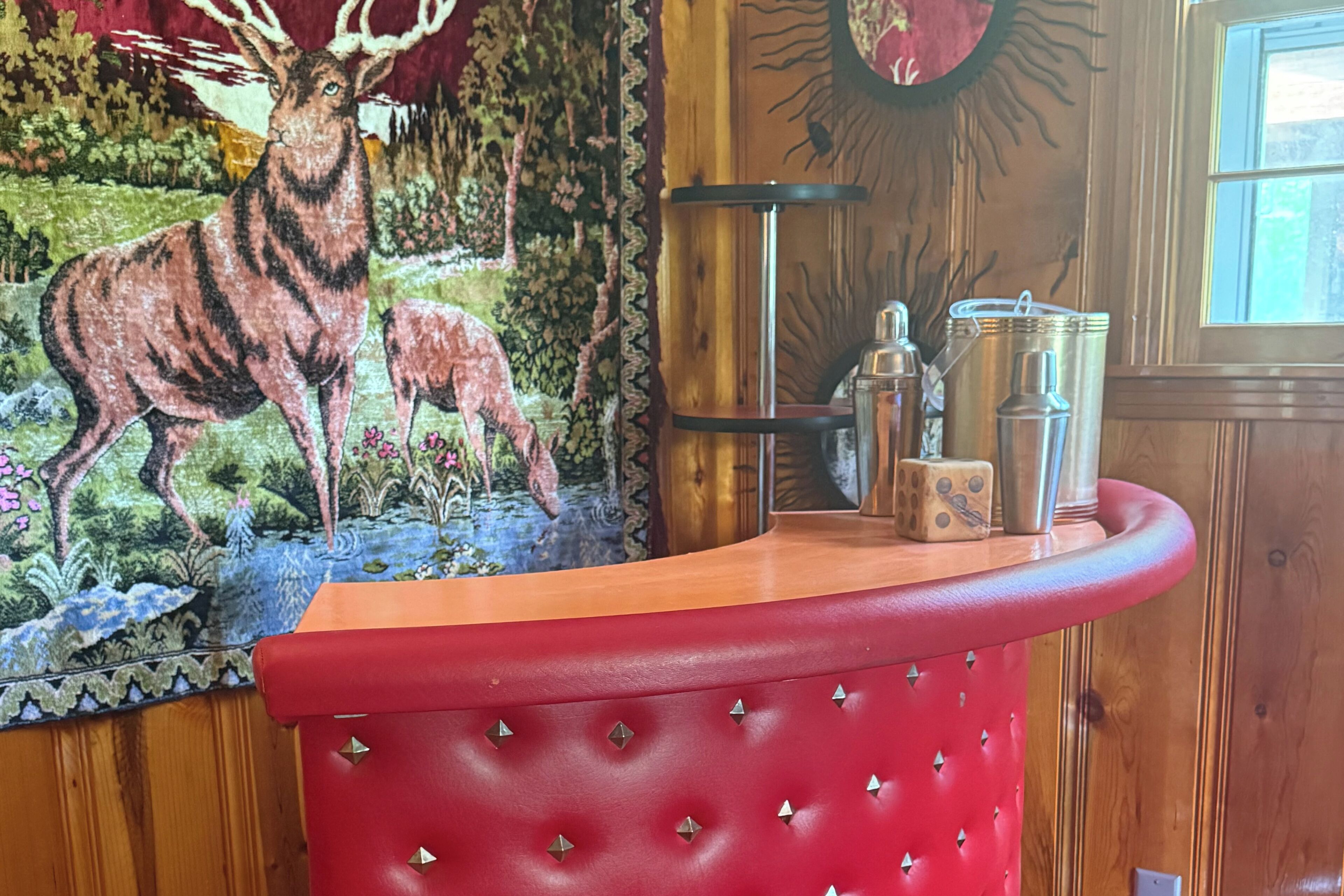 Een rode, beklede bar met een muurschildering van een hert en bos, een metalen barstoel en een raam met natuurlijk licht.