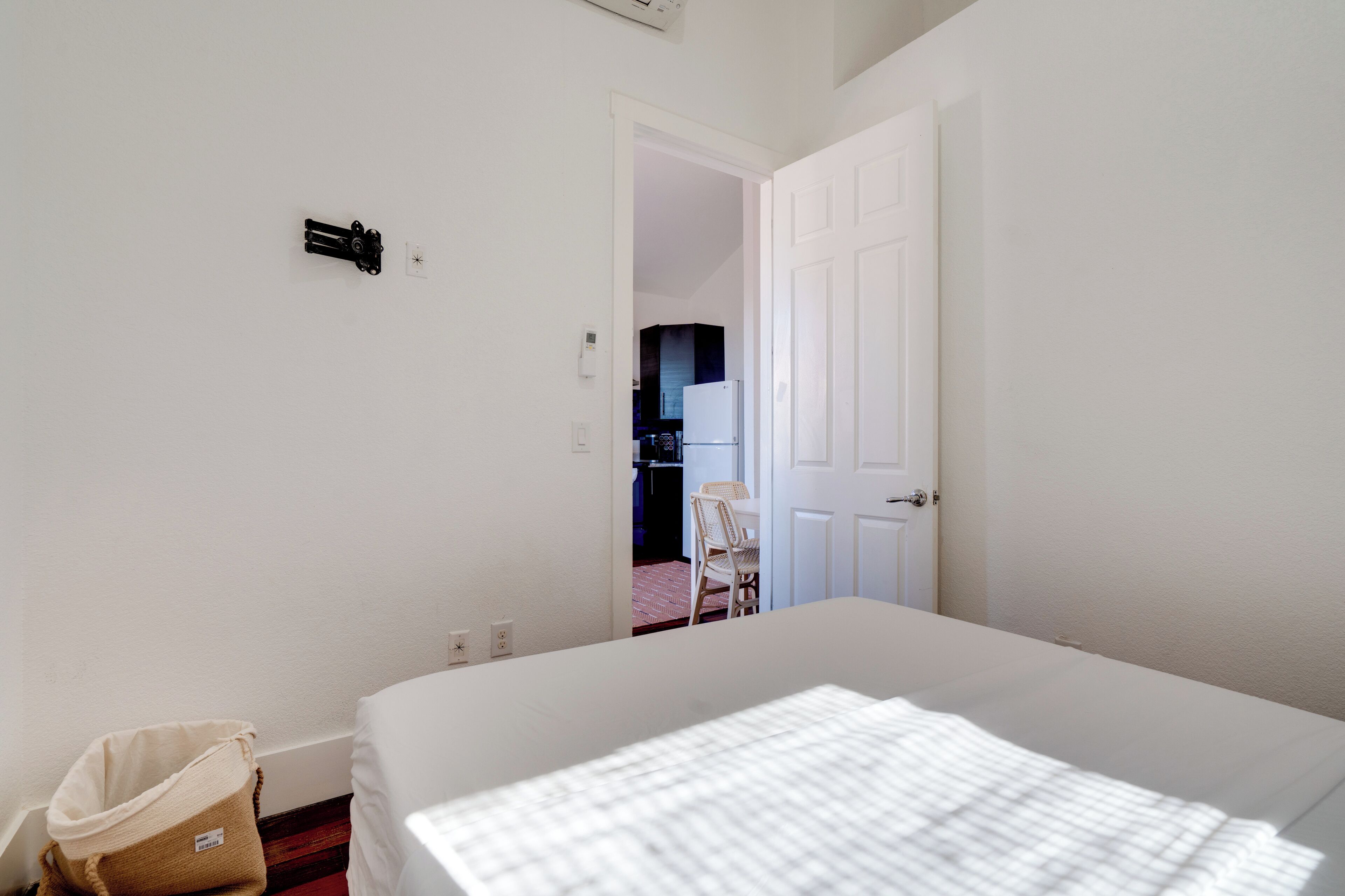 A bedroom with a bed, a white door, and a view into a kitchen area.