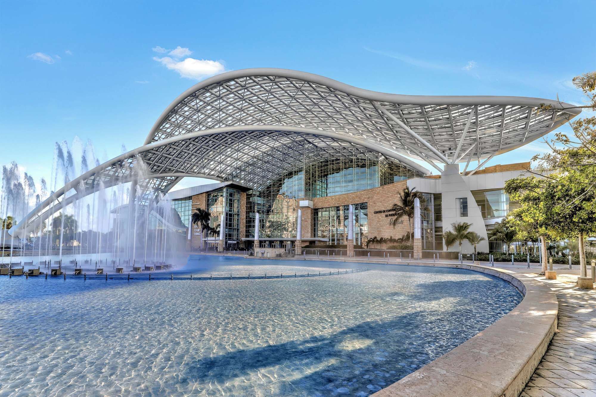 Ein modernes Gebäude mit außergewöhnlicher, gebogener Dachkonstruktion, blauem Himmel und einem Brunnen im Vordergrund.
