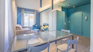 Un espace repas moderne avec une table en verre et des chaises blanches, un canapé et un téléviseur en arrière-plan.