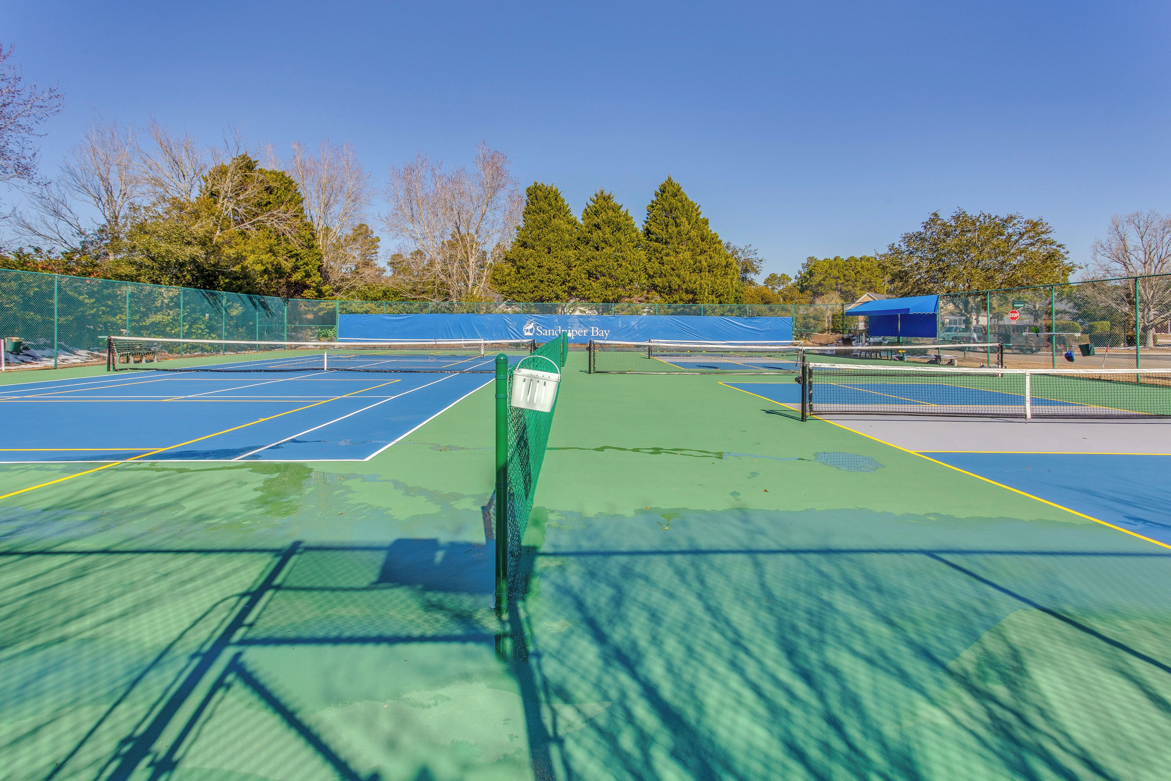 Un court de tennis avec un filet, entouré d’arbres et avec un bâtiment bleu en arrière-plan.