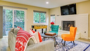 A living room with a sofa, armchair, and a foosball table. There is a fireplace and a TV mounted on the wall.